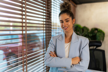 With her arms casually crossed and a warm smile, she stands confidently leaning against her office desk, showcasing her approachable demeanor.