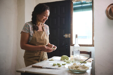 Authentic portrait of multi ethnic young charming woman, housewife in beige apron, standing at kitchen table with ingredients and spinach dough, modeling dumplings in rural kitchen rustic interior