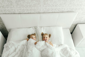 Fototapeta na wymiar Two Little Girls Laying in White Sheet Covered Bed