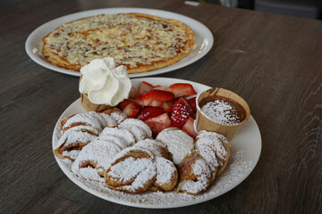 a plate of poffertjes, a traditional Dutch pancake, covered in sugar powder with a side of strawberry fruit and chocolate sauce. A savory pancake with a cheese, bacon, and egg in the background