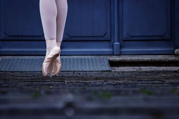 Closeup of a dancer legs balancing on the pointe shoes. Ballerina wearing ballet slippers while standing on her tips on the cobblestone road. Grey retro doors on the background. Selective focus