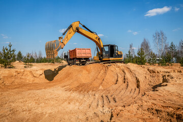 A wheeled excavator loads a dump truck with soil and sand. The excavator digs the ground at the...