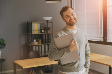Modern happy bearded office male worker of IT company smiling and holding laptop in office