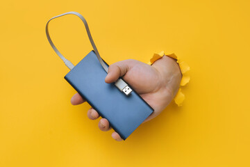 A man's hand with a gray metal power bank protrudes from a torn hole in yellow paper. Concept of smartphone charging and mobile energy independence.