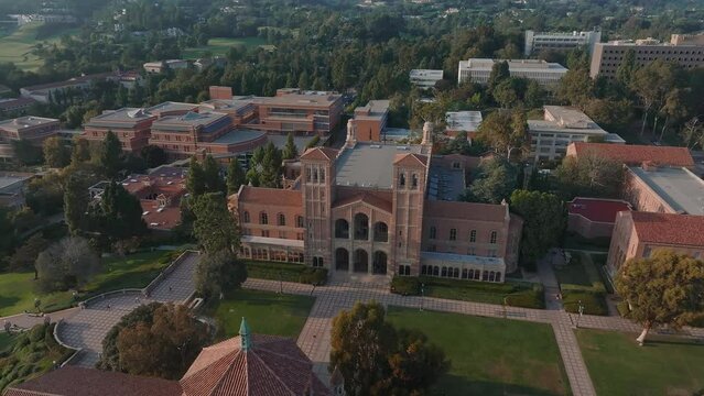 Aerial view of UCLA campus bathed in golden light, showcasing Romanesque Revival and Gothic architecture amid lush greenery, with Royce Hall as the centerpiece.
