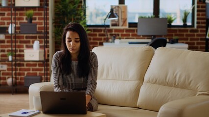 Indian teleworker sitting on couch, focused on finishing tasks in personal office. Remote worker concentrating on correctly inputting data on laptop, hurrying to complete project before deadline