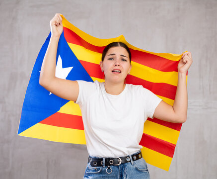Frustrated young woman waving national flag of Estelada while standing against gray wall indoors
