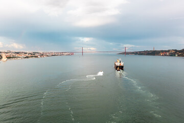 Aerial drone shot of cargo ship moving in Tagus river with Ponte 25 de Abril in background. The iconic Ponte 25 de Abril spans the background, set against the backdrop of a cloudy sky.
