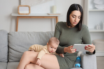 Young woman with tablet computer and her baby suffering from postnatal depression at home