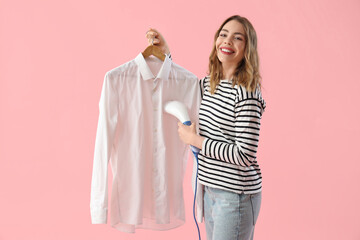 Pretty young woman steaming shirt with modern garment steamer on pink background