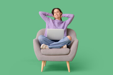 Fototapeta na wymiar Beautiful young happy woman with laptop resting on armchair against green background