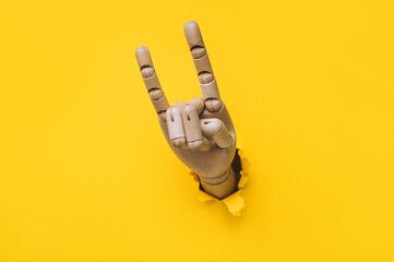 A wooden hand protrudes from a torn hole in yellow paper and shows two fingers, index and little...