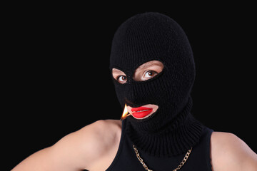 Beautiful young stylish woman in balaclava with match on black background