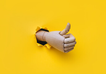 A wooden right hand protrudes from a torn hole in yellow paper and shows a big thumbs up like gesture. The concept of approval and that everything will be fine. Artificial intelligence, robot.