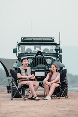 Man and woman laugh joyfully while sitting in camping chairs, with a off-road car in the backdrop, enjoying their outdoor adventure.