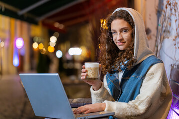Young teenage girl study education online distant job with laptop in city street cafe restaurant browsing website chatting outdoor during break drinking hot coffee. Woman tourist with netbook at night