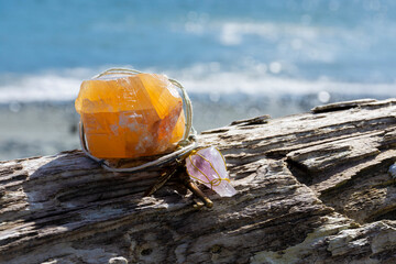 An image of a large orange calcite crystal with handmade pendulum resting on an old piece of driftwood with a blue ocean background. 