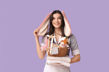 Happy female chambermaid in bunny ears with Easter basket and stack of towels on purple background