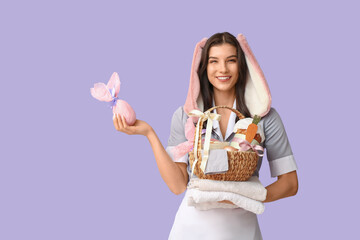 Happy female chambermaid in bunny ears with Easter basket, chocolate egg and towels on purple background