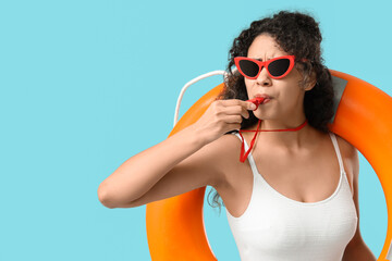 Beautiful young African-American female lifeguard with ring buoy whistling on blue background