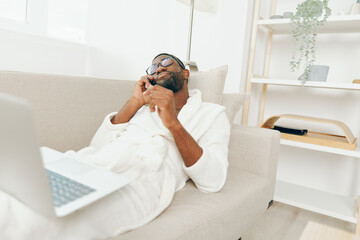 Cheerful African American freelancer in bathrobe sitting on a sofa with a laptop and phone, working...