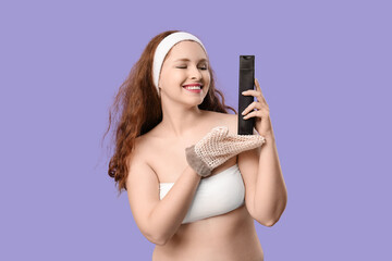 Beautiful young happy woman with bath massage mitten and bottle of shower gel on purple background