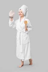 Beautiful young happy woman in bathrobe with bath mitten and massage brush waving hand on grey...