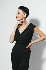 Confident mature woman in stylish black jumpsuit posing with hands on hips for camera in studio...