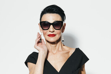 Elegant older woman in black dress and sunglasses posing in front of white wall portrait
