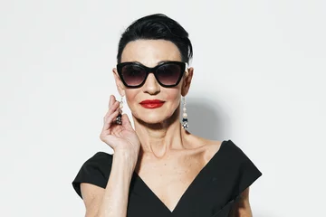 Fotobehang Elegant older woman in black dress and sunglasses posing in front of white wall portrait © SHOTPRIME STUDIO