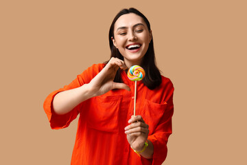 Beautiful young woman with sweet lollipop showing half heart gesture on brown background