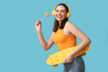 Beautiful young woman in headphones with sweet lollipop and skateboard on blue background