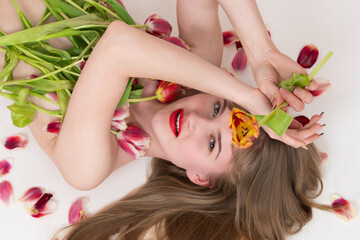 Obraz na płótnie Canvas Half naked blonde woman is lying on white background with bouquet of spring tulips for Valentine's Day covering her chest. Sensuality young Caucasian blonde woman looking at camera in romantic pose