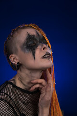 Informal young woman with orange color dreadlocks hairstyle and creative horror black stage makeup painted on face. Studio shot on blue background. Part of photo series