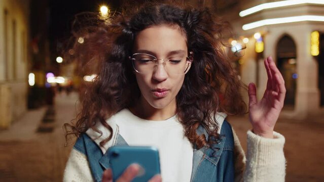 Oh my God Wow. Excited happy joyful winner pretty young woman using smartphone typing browsing celebrating win good message news. Girl with brown windswept hair on urban city street at night evening.