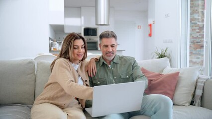 Happy middle aged couple using laptop relaxing on couch at home. Smiling mature man and woman looking at computer watching video, browsing or shopping in ecommerce store sitting on sofa in living room - Powered by Adobe