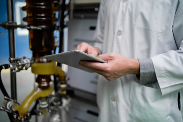 A scientist wearing a lab coat and goggles is writing on a clipboard. The clipboard is black and the scientist is looking at it intently