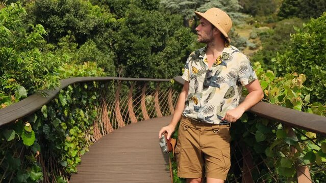 a male traveler in a safari hat takes a photo on a suspension bridge. traveler with backpacks relaxing in greens jungle and enjoying view in waterfall. tourist traveling along rocky forest trek.