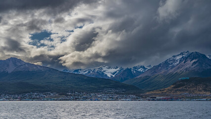 Beautiful snow-capped mountain range of the Andes against the sky and clouds. The city of Ushuaia is at the foot of the mountains, on the banks of the Beagle Canal. Ripples on the blue water.