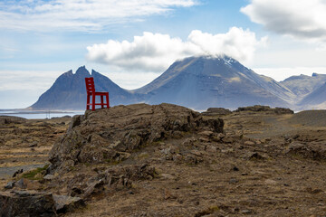 A red chair is perched on a rocky hill overlooking a stunning natural landscape of mountains, hills, and a cloudy sky in the background. Iceland