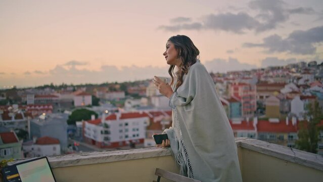 Serene woman peeping cityscape balcony with coffee cup in hands. Lady waiting