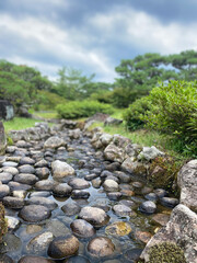 Fototapeta na wymiar A tranquil pebble-filled stream bed with clear water flowing among the rounded stones. Lush greenery surrounds the stream under a partly cloudy sky.