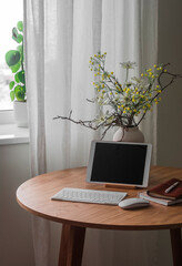 A tablet, a keyboard, a vase of flowers on a round wooden table near the window in the living room