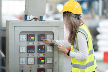 Asian female engineer with long hair pressing an electronic control panel controlling a system within an industrial factory, logistics business, holding a tablet. Wear a safety helmet and vest.