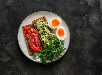 Rye crackers with avocado and salmon, boiled egg, arugula - delicious breakfast, snack on a dark background, top view