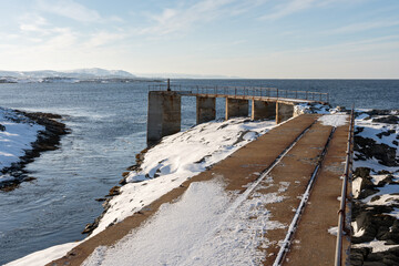A concrete bridge by the Kjolnes Lighthouse by the Barents Sea on a sunny winter day in March, town of Berlevåg in the background, Norway