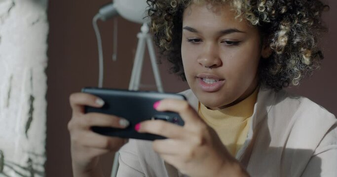 Joyful African American woman playing online video game with smartphone relaxing in office in work break. Entertainment and gaming device concept.