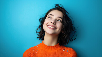 Portrait of a happy, young girl with dark hair wearing an orange sweater, looking upwards with a joyful expression..