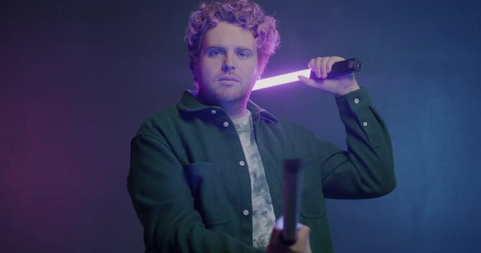 Slow motion portrait of creative young man moving hands with neon light sticks and looking at camera on purple background. People and illumination concept.