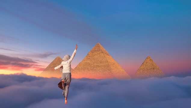 Muslim woman in gray trousers and a scarf jumps on the sands of the Sahara desert -  Giza Pyramid Complex at amazing sunset - Cairo, Egypt 